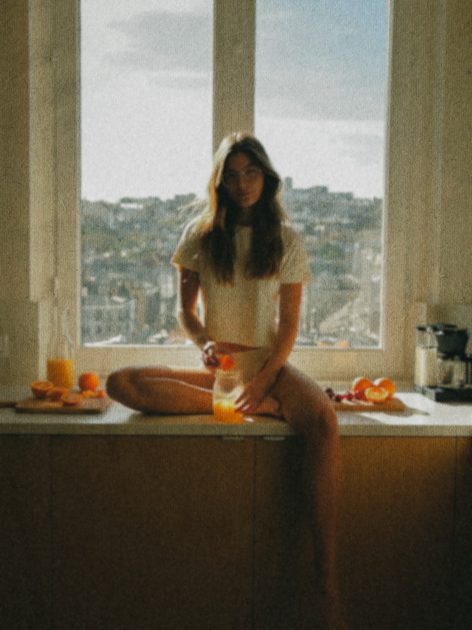 Person sitting on a windowsill with a cityscape view, holding a drink.